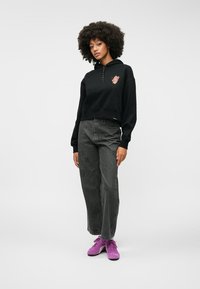 Czarna krótsza bluza z kapturem z grafiką na piersi, w zestawieniu z szarymi dżinsami o wysokim stanie i różowymi trampkami.