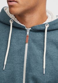 Kapuce s zipem v barvě teal, s měkkou fleecovou podšívkou. Mezi detaily patří kovový zip, textilní stahovací šňůrky a kožený štítek s logem.
