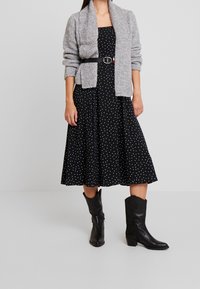 Svart klänning med prickar i A-linjeform, kombinerad med en grå texturerad cardigan och svarta lädercowboyboots. Bälte vid midjan.