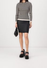 Kvinna iförd en svart och vit randig långärmad topp, svart mini kjol, svarta platåskor, hållande en svart handväska mot vit bakgrund.