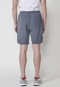 Person som står vänd bortåt iklädd texturerade grå shorts, vita sneakers med röda och navy accenter, samt en vit shirt.