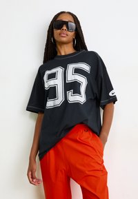 Svart oversized t-skjorte med hvit trykk av nummer "95", korte ermer og rund hals, kombinert med oransje nylonbukser.
