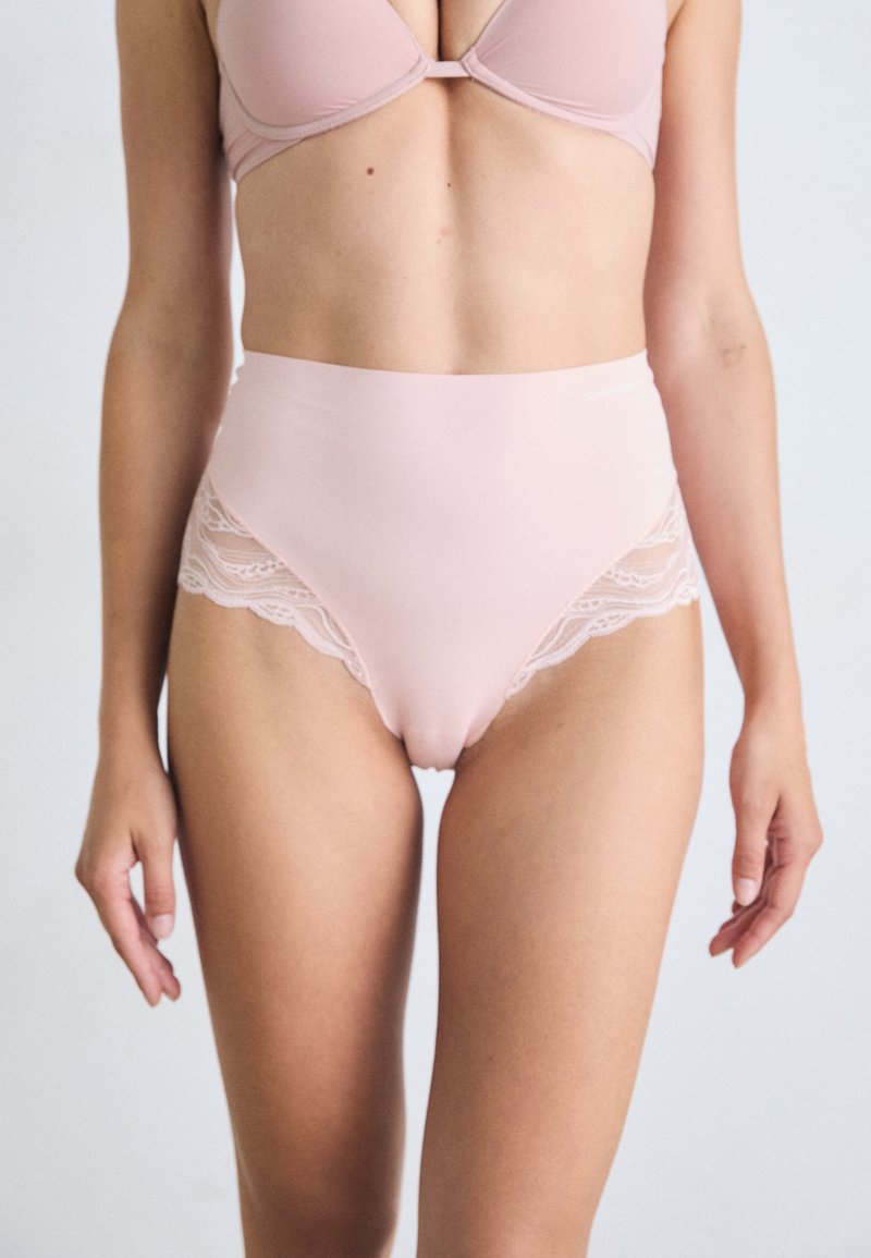 Mutande alte di colore rosa chiaro, realizzate in un tessuto liscio con bordo in pizzo lungo le aperture delle gambe, che offrono un delicato contrasto di texture.