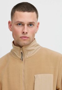 Tan fleece jacket with high collar, front zipper, and side pocket. Smooth texture and simple design, suitable for casual wear.