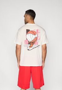 Μπεζ κοντομάνικη μπλούζα με γραφικό που απεικονίζει ένα χέρι να κρατά ένα μπάσκετ, με ένα πολύχρωμο αφηρημένο φόντο. Συνδυάζεται με κόκκινα σορτς.