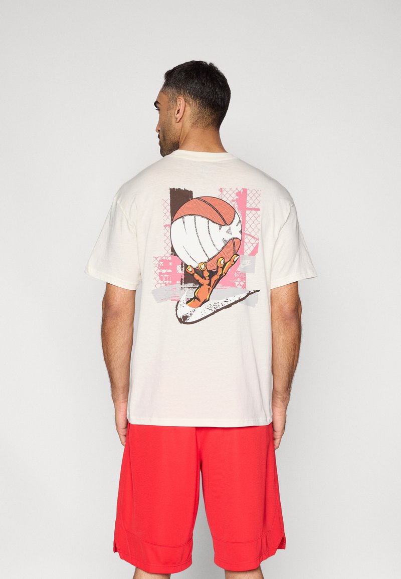 Μπεζ κοντομάνικη μπλούζα με γραφικό που απεικονίζει ένα χέρι να κρατά ένα μπάσκετ, με ένα πολύχρωμο αφηρημένο φόντο. Συνδυάζεται με κόκκινα σορτς.