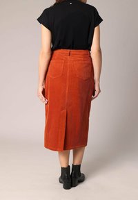 Jupe midi en velours côtelé rouille avec une fente sur le côté, dotée de deux poches arrières. Associée à des bottines noires et un haut noir à manches courtes.
