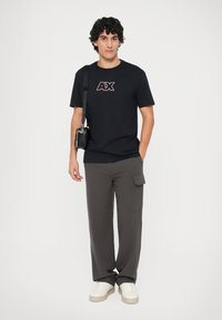 Camiseta azul marino con un logo "AX" rojo y blanco, combinada con pantalones cargo de pierna ancha en gris. El modelo sostiene un pequeño bolso negro y lleva zapatillas blancas.