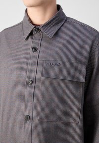 Button-up shirt in a check pattern, featuring shades of blue, grey, and burgundy, with a chest pocket and a collared neckline.