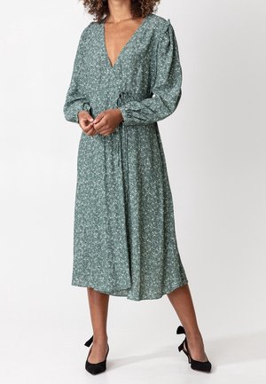 Femme portant une robe midi fleurie verte à manches longues et des talons noirs, debout devant un fond uni.