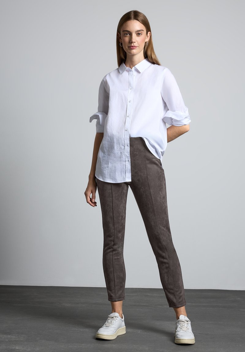 Witte button-up shirt met opgerolde mouwen, gecombineerd met grijze suede-achtige broek en witte sneakers. Gladde texturen, aansluitend ontwerp en minimale details.