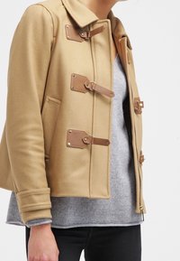 Vrouw in een beige wollen jas met leren gespbanden over een grijze trui en zwarte broek, tegen een effen witte achtergrond.