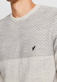 Maglione grigio a maglia con texture, caratterizzato da un motivo a rombi nella parte superiore e una parte inferiore liscia. Presenta un piccolo logo di un uccellino nero sul petto.
