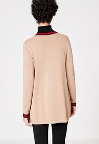 Osoba ubrana w beżowy sweter z długim rękawem, z czarnymi i czerwonymi paskami na mankietach i kołnierzu, stojąca tyłem na jednolitym tle.