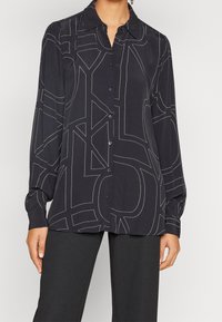 Blusa nera con bottoni e maniche lunghe. Presenta motivi di cucito geometrici bianchi. Realizzata in tessuto liscio con colletto strutturato.
