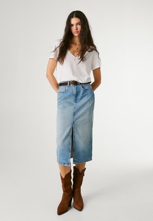 Ung kvinde med langt hår iført hvid V-hals T-shirt, lyseblå denimnederdel med bælte og forreste slids samt brune ruskindsstøvler til anklerne.