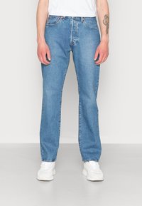 Jeans retos de tom azul claro feitos de denim, com costuras laranja, cinco bolsos e uma cintura padrão, combinados com sapatilhas brancas.