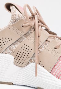 Beżowy but sportowy z teksturowanym dzianinowym wierzchem, gumową podeszwą i różowymi akcentami. Posiada perforowane syntetyczne nakładki i płaskie sznurówki.
