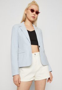 Prugasta blazerica v svetlo modri in beli barvi, z enim gumbom, zarezanimi reverji in dvema sprednjima žepoma, oblečena čez črn crop top.
