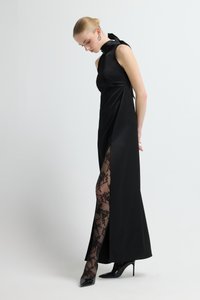 Czarna satynowa suknia z wysokim dekoltem i bocznym rozcięciem, zestawiona z przezroczystymi czarnymi koronkowymi rajstopami i szpilkami z ostro zakończonym noskiem. Gładka tekstura i elegancka sylwetka.