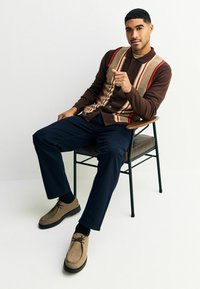 Barna cardigan, vertikális krém és piros csíkokkal, sötét ing fölött viselve; a megjelenést tengerészkék nadrág és világosbarna cipő egészíti ki.