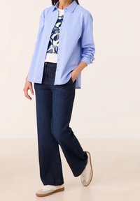 Camicia a righe azzurro chiaro con bottoni, t-shirt bianca con grafica, jeans larghi scuri e sneakers bianche con accenti beige. Design semplice, vestibilità casual.
