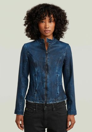 Frau trägt eine taillierte blaue Jeansjacke mit Reißverschluss und Stehkragen sowie schwarze Hose, steht vor einem schlichten hellen Hintergrund.