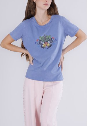 Mujer con camiseta azul con diseño de mariposa y flores, combinada con pantalones rosa claro con adornos laterales.