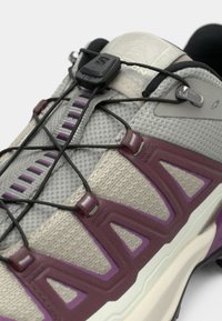 Gros plan d'une basket beige et violette avec un système de laçage rapide élastique noir et une tige en mesh texturé.