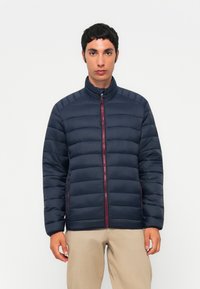 Marineblå pufferjakke med vandret quiltet design, høj krave, frontlynlås med røde detaljer og sidelommer. Letvægtsstof.