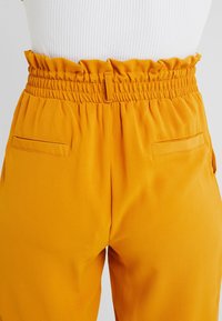 Fitted yellow pants with ruffled elastic waistband, two back pockets, smooth texture, and tapered leg design.