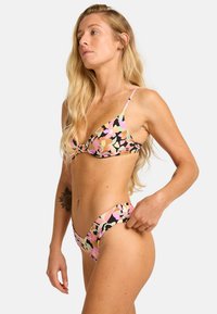 Floral print bikini in shades of pink, green, and black. Features a triangular bra top and low-rise bottoms with minimal coverage and thin straps.