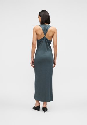 Femme portant une robe maxi sans manches et dos nu de couleur vert canard foncé avec un fermoir doré sur le haut du dos et des mules à talons noires, debout de dos.