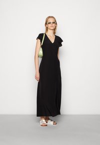 Vestido maxi preto, com botões e mangas curtas em babados. Combinado com sandálias brancas e uma pequena bolsa verde. Textura suave.