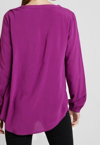 Långärmad, lila blus med rundad halsringning. Slätt, lättviktigt tyg med en rundad fåll och rynkade ärmslut. Svarta byxor.