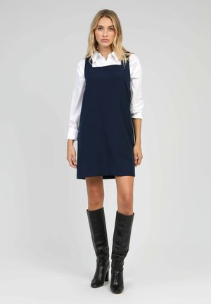 Robe sans manches bleu marine avec décolleté carré, associée à une chemise blanche et des bottes en cuir noir jusqu'aux genoux. Tissu lisse et design simple.