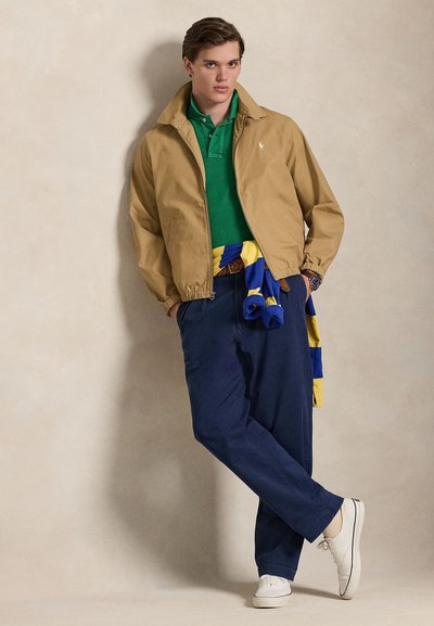 Καφέ μπουφάν bomber, πράσινο πόλο, μπλε σκούρο παντελόνι, λευκά αθλητικά παπούτσια· πολύχρωμο φουλάρι δεμένο γύρω από τη μέση· χαλαρή στάση μπροστά σε φωτεινό φόντο.