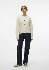 Off-white gebreide cardigan met een ronde hals, knopen aan de voorkant en lange mouwen, gecombineerd met donkere flared jeans en casual schoenen.