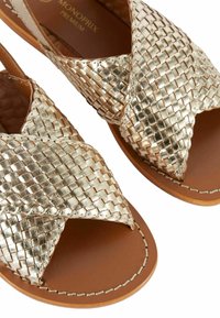 Sandales en or tissées avec des lanières croisées, semelle intérieure marron et semelle plate. La texture est lisse et brillante, mettant en valeur un motif tissé distinctif.