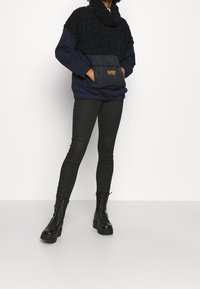 Pullover navy con sezione superiore pelosa, tasca frontale e collo a imbuto; abbinato a pantaloni neri aderenti e stivali neri chunky.