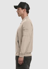 Beige bomberjakke med lynlås foran, ribbede ærmer og en nyrepung på ærmet. Glat tekstur og letvægtsmateriale.