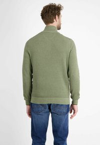 Grüner gestrickter Pullover mit hohem Kragen, gerippten Bündchen und Saum, der ein strukturiertes Muster aufweist. Getragen mit blauen Jeans, von hinten betrachtet.