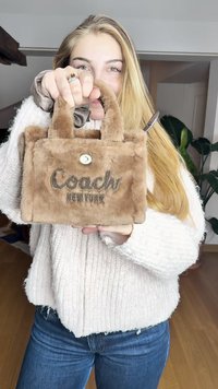 Καφέ τσάντα από ψεύτικη γούνα με κοντούς χειρολαβές, που διαθέτει το «Coach New York» ραμμένο με σκούρο νήμα και κυκλικό μεταλλικό κούμπωμα.