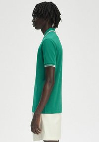 Polo vert avec col et bord de manches blancs, à manches courtes et coupe droite. Texture lisse, porté avec un short de couleur claire.