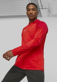 Rote Sportjacke mit strukturierten Ärmeln, die ein geometrisches Muster aufweisen, hohem Kragen, durchgehendem Reißverschluss und Logo auf der Brust.