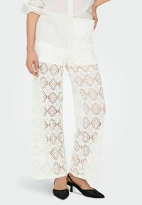 Pantaloni a gamba larga in pizzo bianco con intricati motivi a diamante, caratterizzati da una vita alta e una texture trasparente. Abbinati a scarpe nere con tacco.