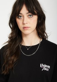 Czarna blouse z długim rękawem z logo w kolorze purpurowym i białym. Modelka nosi nałożone na siebie srebrne naszyjniki z prostym wisiorkiem.