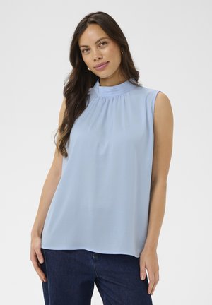 Femme aux longs cheveux foncés portant un chemisier sans manches bleu clair à col haut et un jean bleu foncé, debout devant un fond uni.