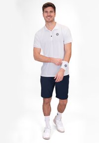 Weißes kurzärmliges Poloshirt mit Kragen und Logo, kombiniert mit marineblauen Shorts. Das Modell trägt weiße Sportschuhe und Knöchelsocken.