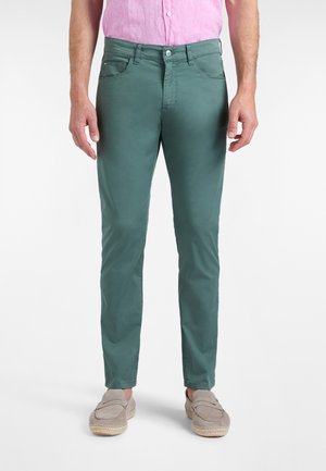 Mand iført slanke grønne bukser, lyserød skjorte med knapper og lysegrå slip-on sko, stående mod en hvid baggrund.